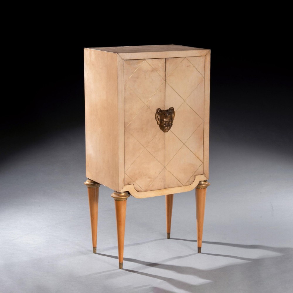 A cream parchment-veneered Art Deco standing cabinet featuring diamond-incised door panels, a central bronze mask handle, and slender tapered legs.