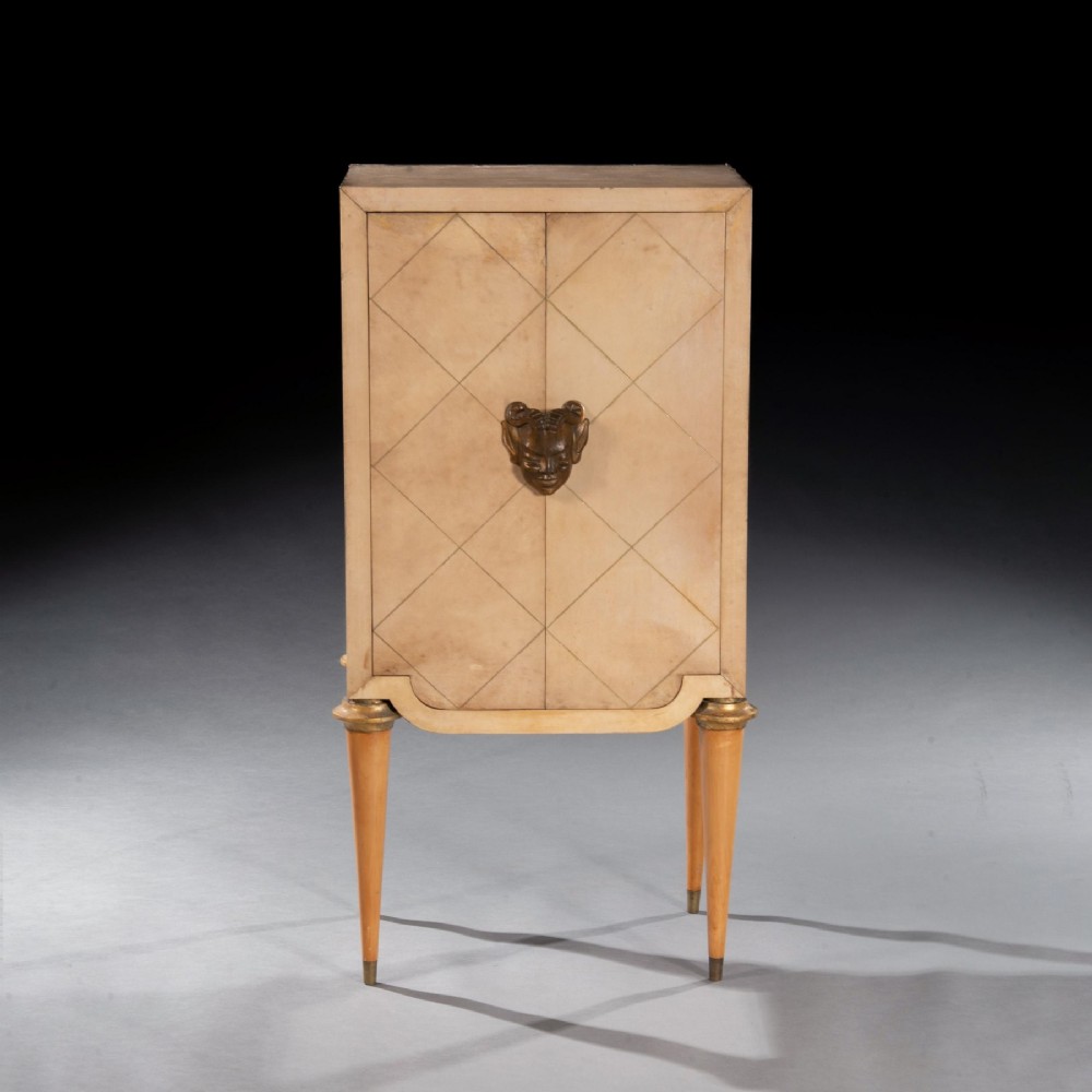 A cream parchment-veneered Art Deco standing cabinet featuring diamond-incised door panels, a central bronze mask handle, and slender tapered legs. View II