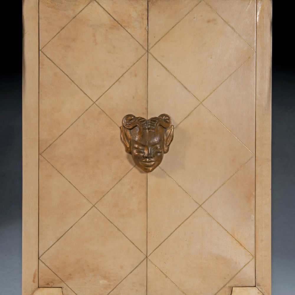 A cream parchment-veneered Art Deco standing cabinet featuring diamond-incised door panels, a central bronze mask handle, and slender tapered legs. View III