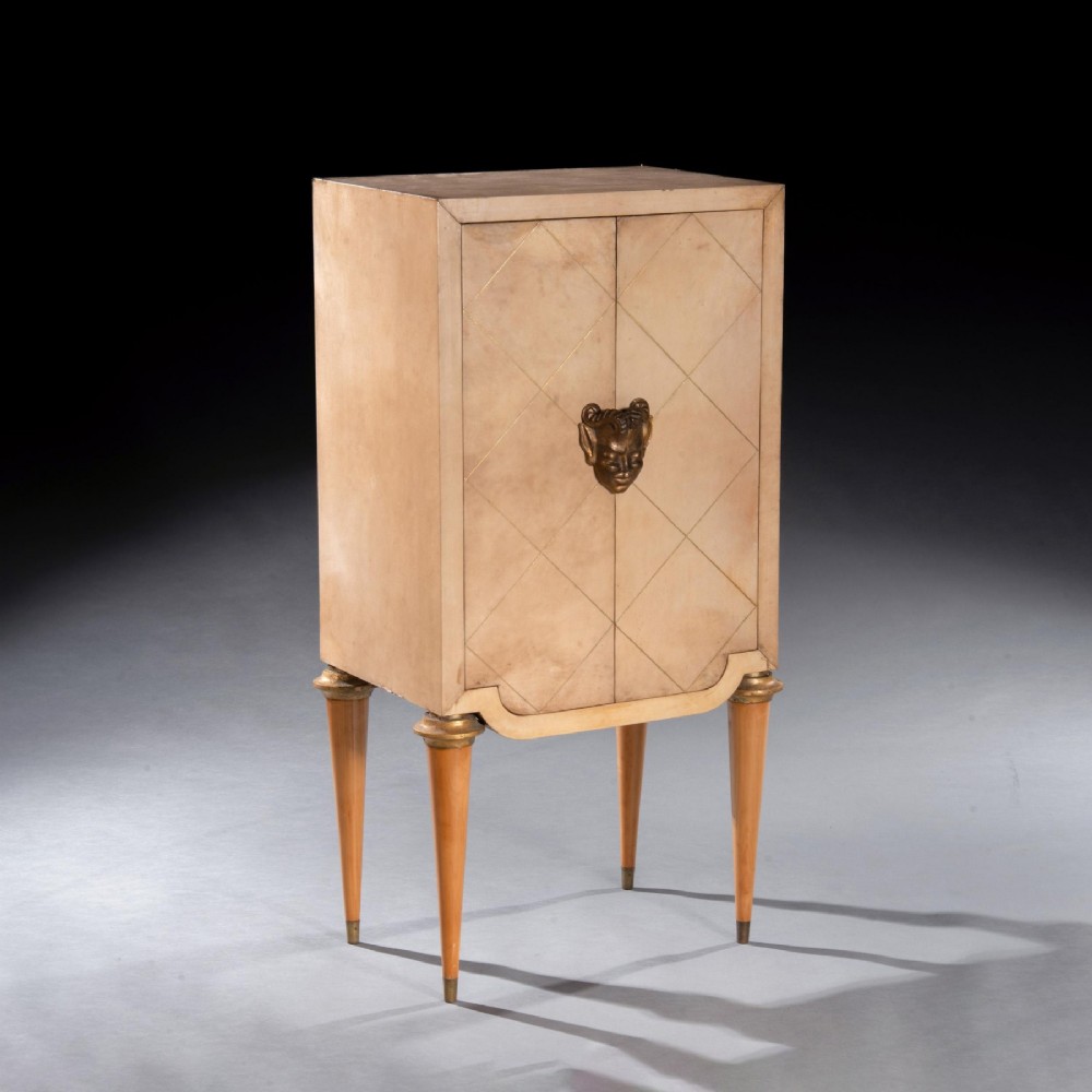 A cream parchment-veneered Art Deco standing cabinet featuring diamond-incised door panels, a central bronze mask handle, and slender tapered legs. View VI