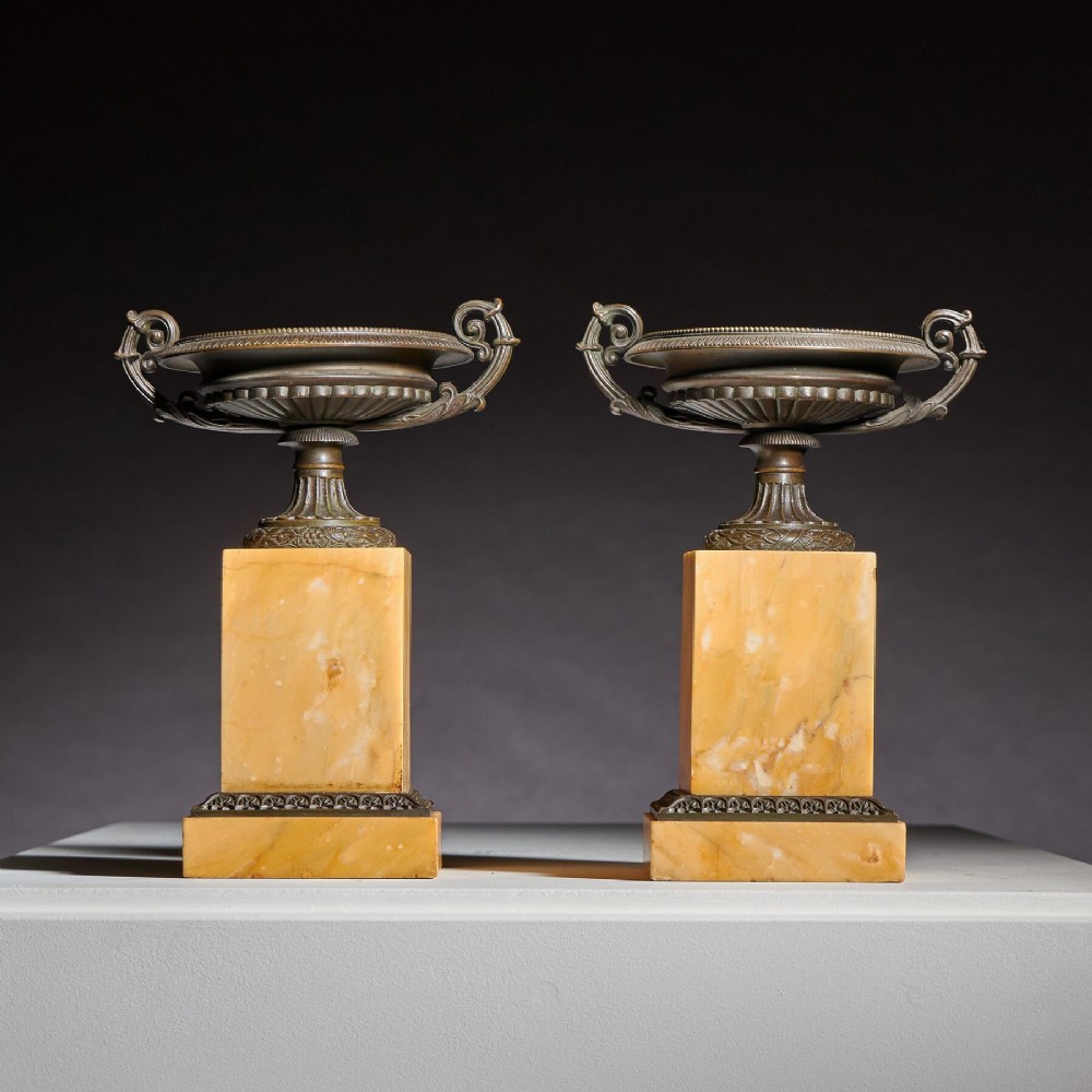 A pair of early 19th-century bronze tazzas with scroll handles mounted upon tall rectangular yellow Siena marble plinths.