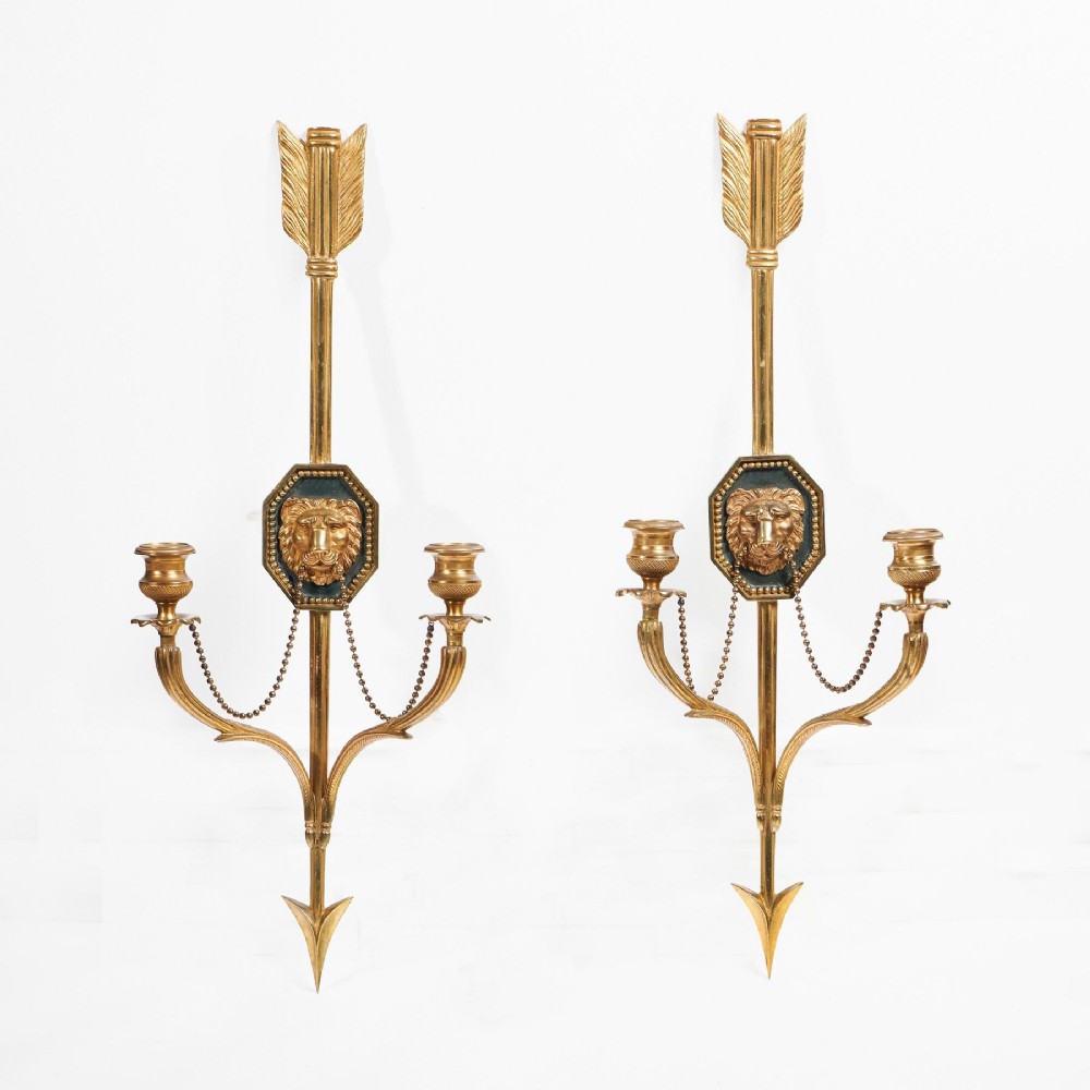 A pair of vertical gilt-metal two-light wall appliques shaped like arrows, featuring central dark octagonal medallions with lion mask reliefs and draped chains.