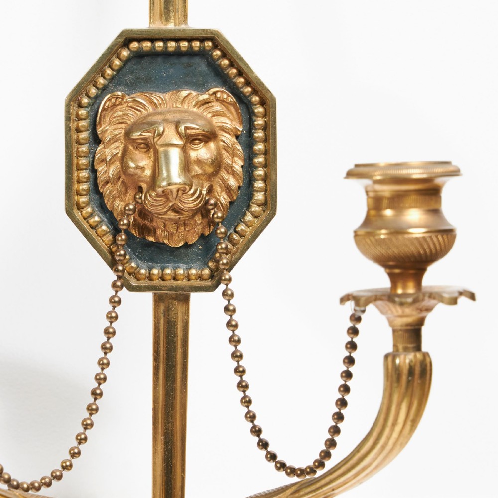 A pair of vertical gilt-metal two-light wall appliques shaped like arrows, featuring central dark octagonal medallions with lion mask reliefs and draped chains. View II