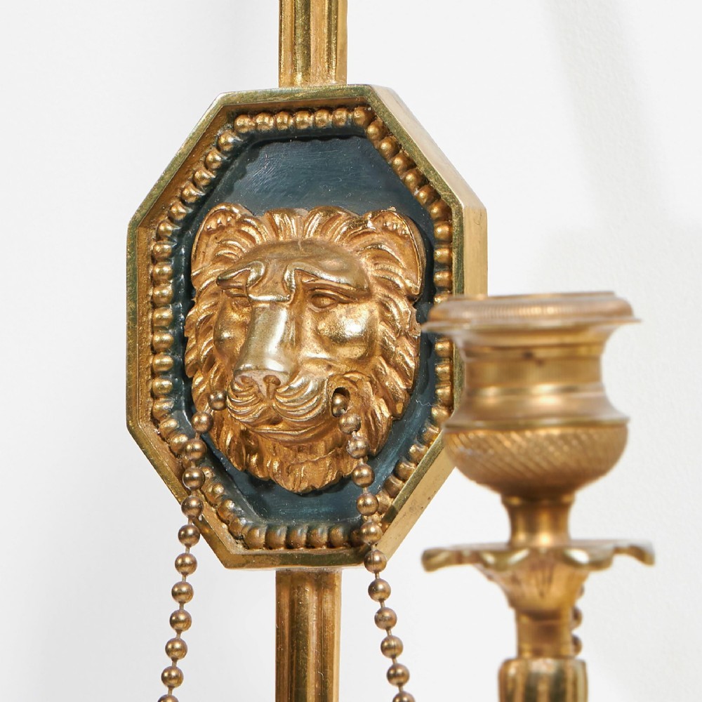 A pair of vertical gilt-metal two-light wall appliques shaped like arrows, featuring central dark octagonal medallions with lion mask reliefs and draped chains. View III