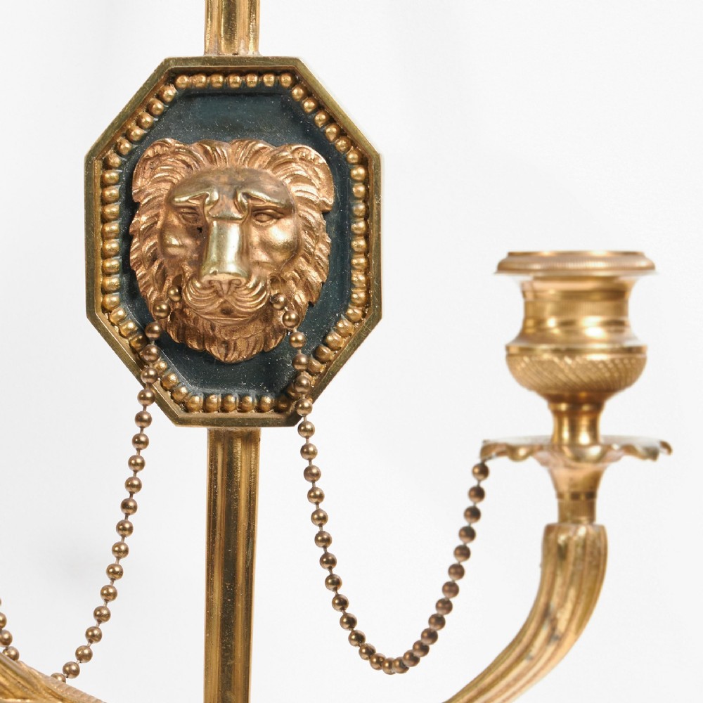 A pair of vertical gilt-metal two-light wall appliques shaped like arrows, featuring central dark octagonal medallions with lion mask reliefs and draped chains. View V