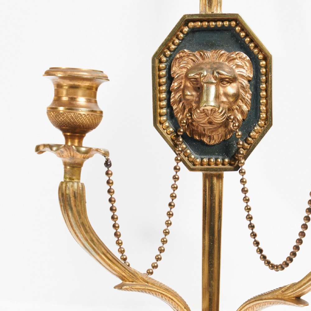 A pair of vertical gilt-metal two-light wall appliques shaped like arrows, featuring central dark octagonal medallions with lion mask reliefs and draped chains. View VI