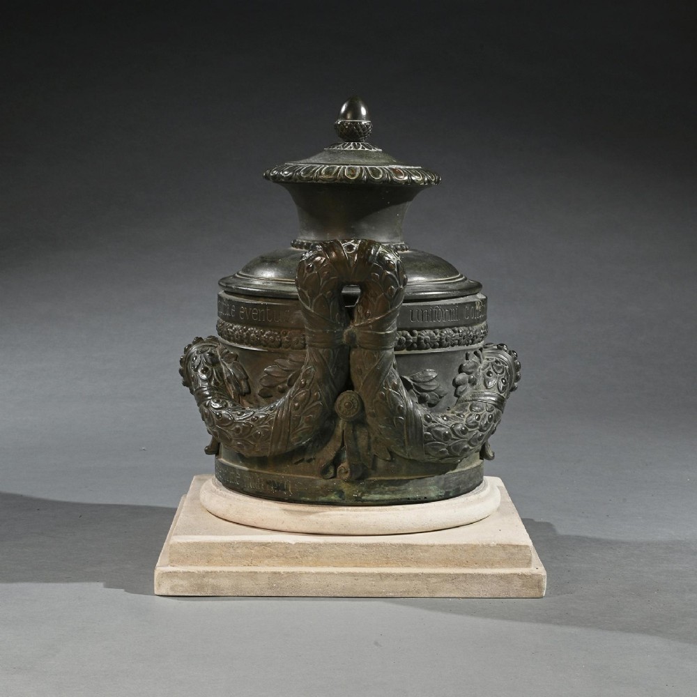 A dark patinated 18th-century Italian bronze vessel featuring relief garlands, handles, and Latin inscriptions, resting on a beige stepped stone plinth. View V
