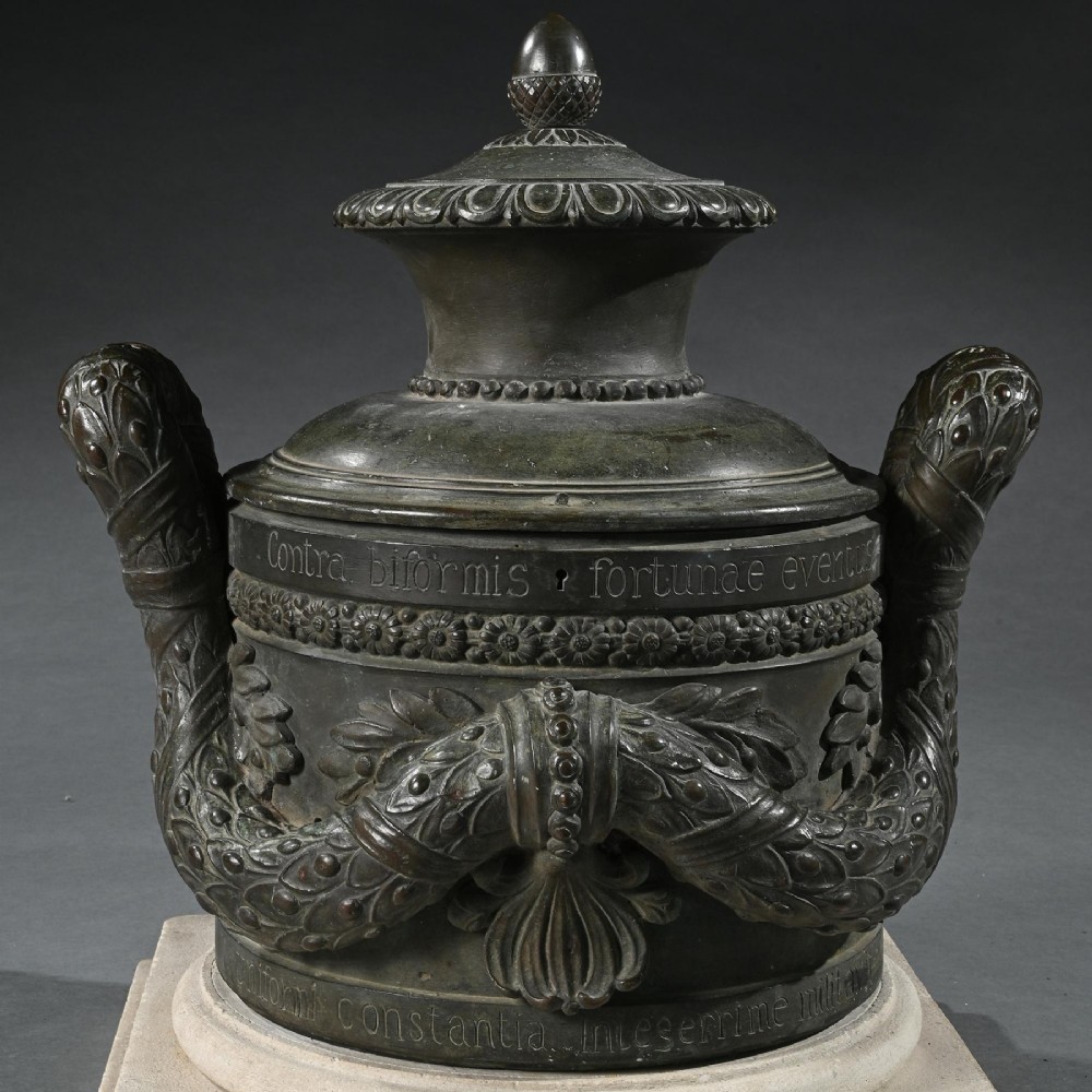 A dark patinated 18th-century Italian bronze vessel featuring relief garlands, handles, and Latin inscriptions, resting on a beige stepped stone plinth. View VI