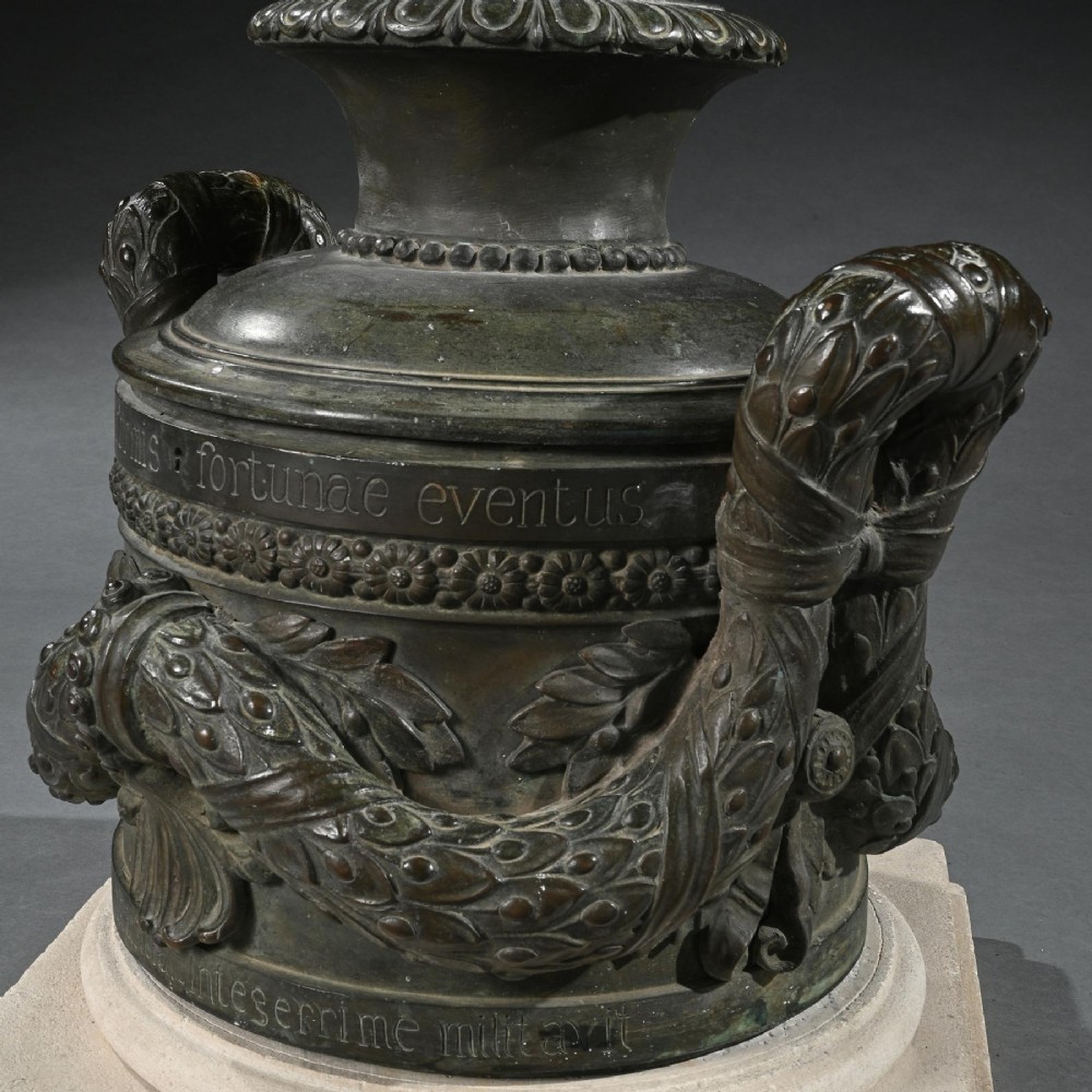 A dark patinated 18th-century Italian bronze vessel featuring relief garlands, handles, and Latin inscriptions, resting on a beige stepped stone plinth. View VII