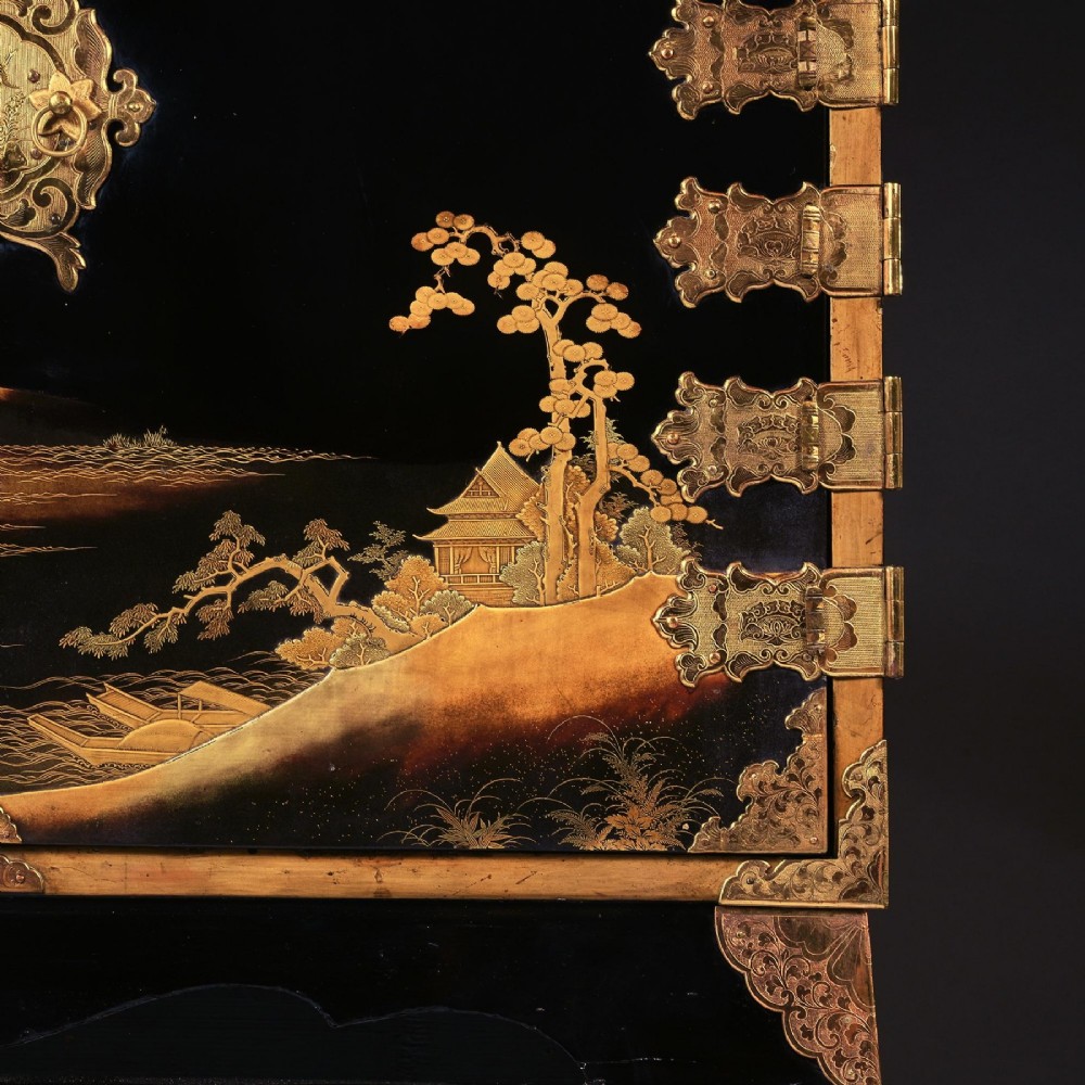 A black lacquered Japanese cabinet decorated with gold maki-e landscape scenes, featuring ornate brass mounts and elevated on a simple black stand. View V
