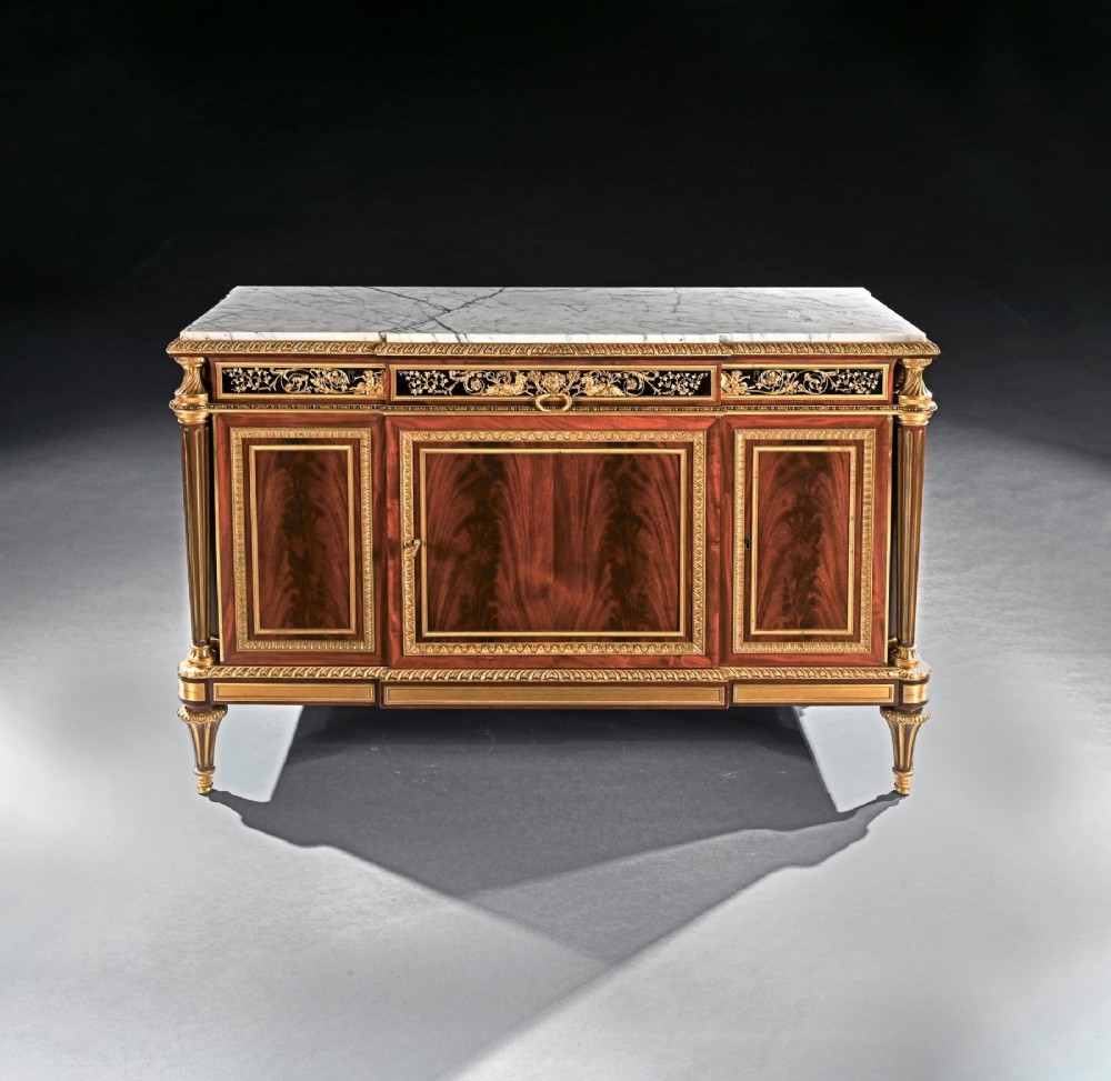 A 19th-century French Louis XVI style flame mahogany commode featuring ornate gilt-bronze mounts, three paneled cupboard doors, fluted legs, and a white marble top. View II