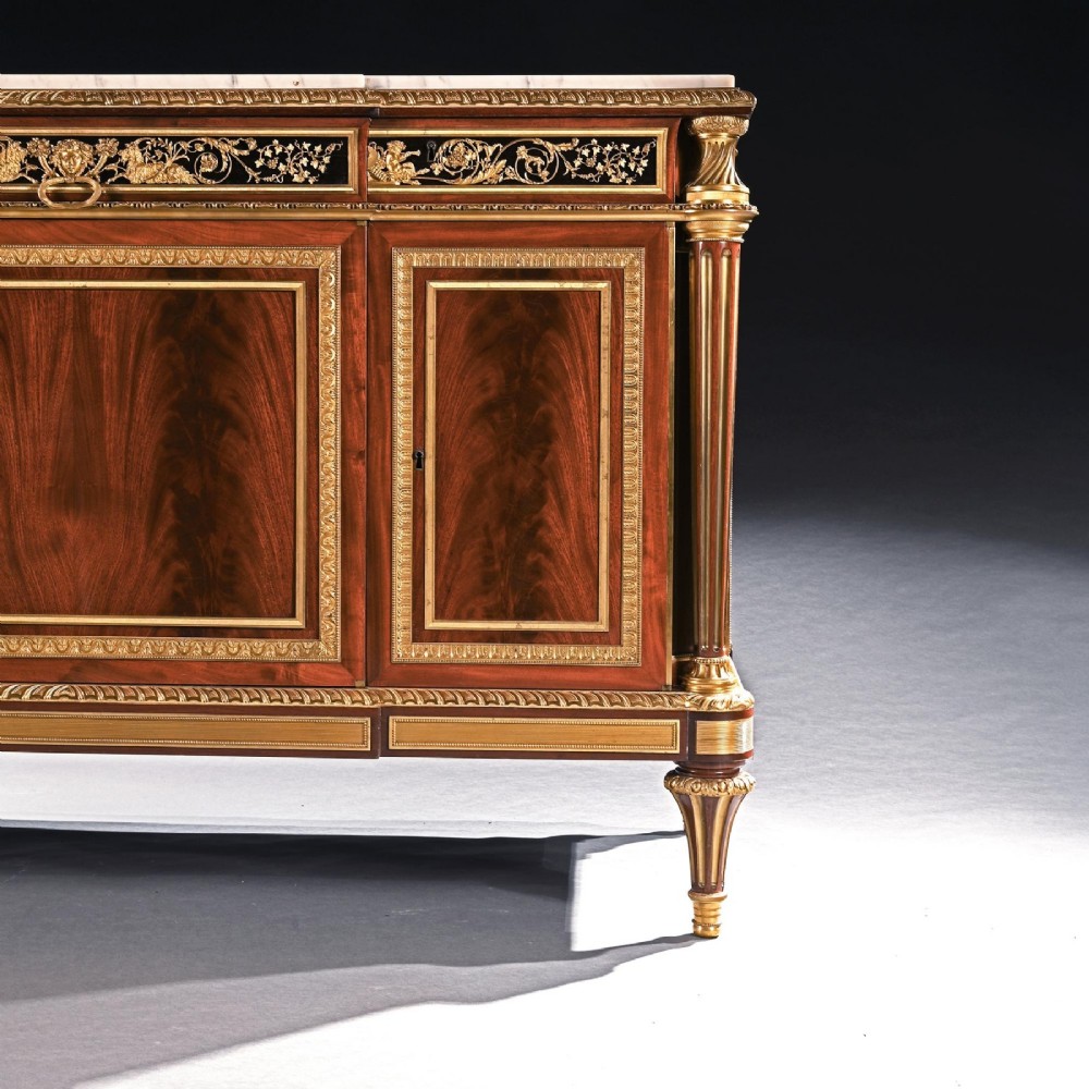 A 19th-century French Louis XVI style flame mahogany commode featuring ornate gilt-bronze mounts, three paneled cupboard doors, fluted legs, and a white marble top. View IV