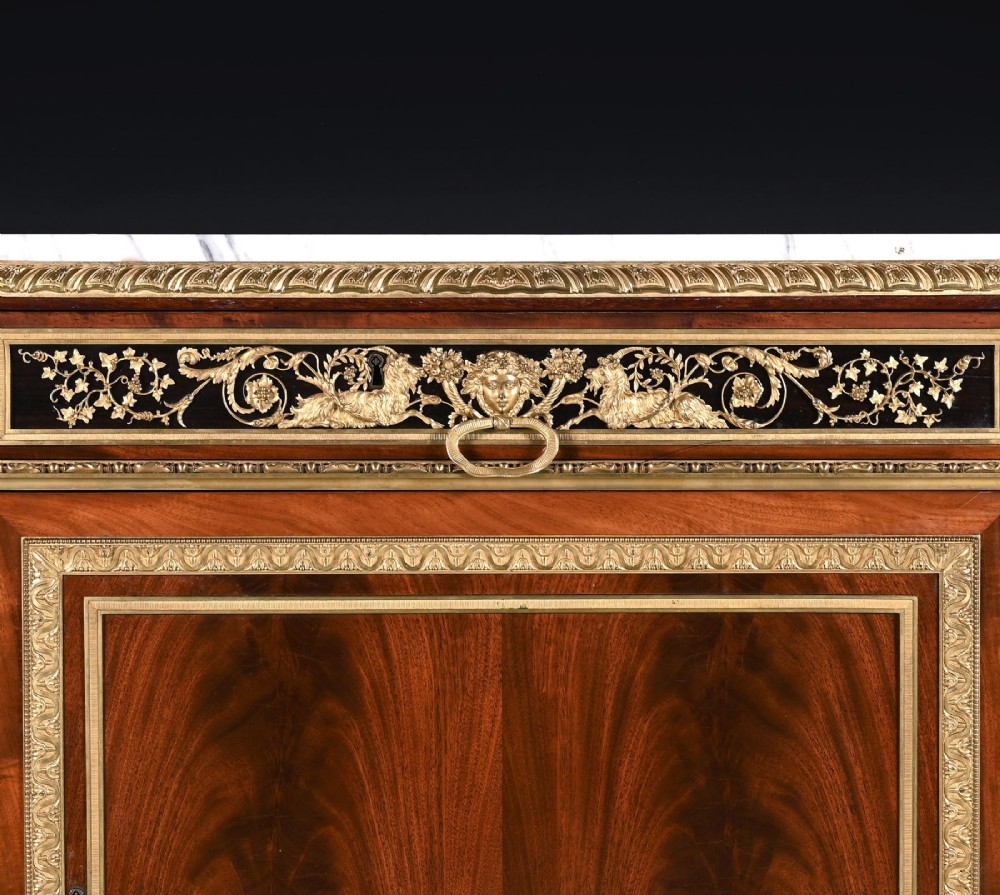 A 19th-century French Louis XVI style flame mahogany commode featuring ornate gilt-bronze mounts, three paneled cupboard doors, fluted legs, and a white marble top. View V