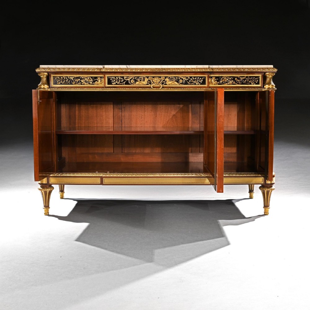 A 19th-century French Louis XVI style flame mahogany commode featuring ornate gilt-bronze mounts, three paneled cupboard doors, fluted legs, and a white marble top. View VI