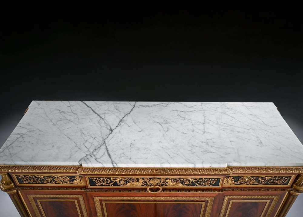 A 19th-century French Louis XVI style flame mahogany commode featuring ornate gilt-bronze mounts, three paneled cupboard doors, fluted legs, and a white marble top. View VII