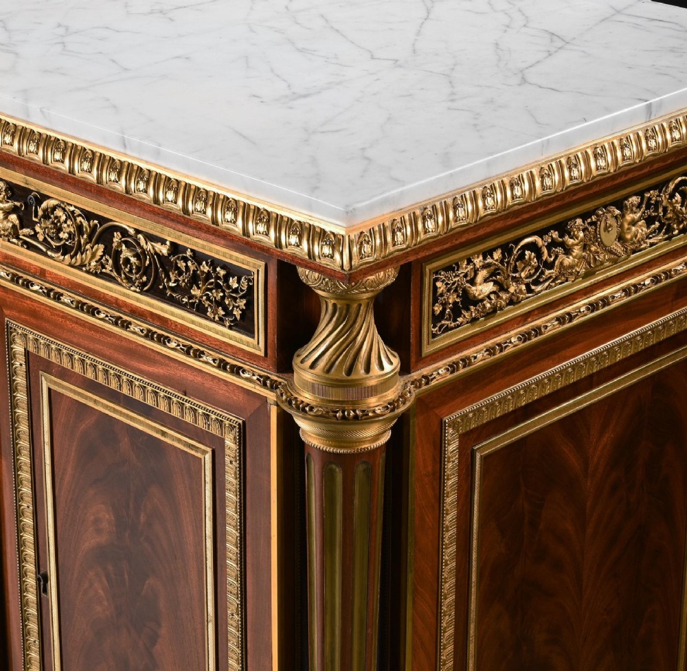A 19th-century French Louis XVI style flame mahogany commode featuring ornate gilt-bronze mounts, three paneled cupboard doors, fluted legs, and a white marble top. View VIII