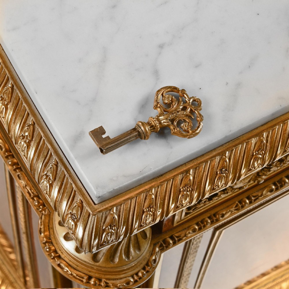 A 19th-century French Louis XVI style flame mahogany commode featuring ornate gilt-bronze mounts, three paneled cupboard doors, fluted legs, and a white marble top. View IX