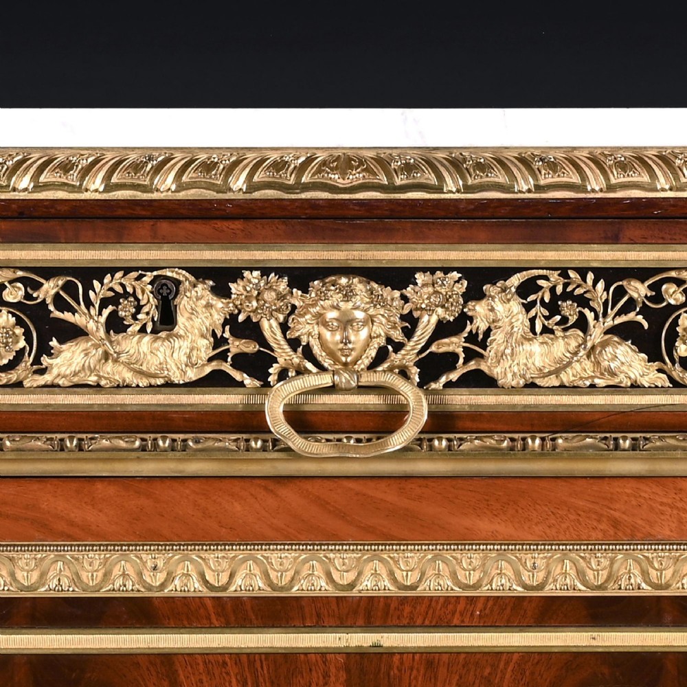 A 19th-century French Louis XVI style flame mahogany commode featuring ornate gilt-bronze mounts, three paneled cupboard doors, fluted legs, and a white marble top. View X