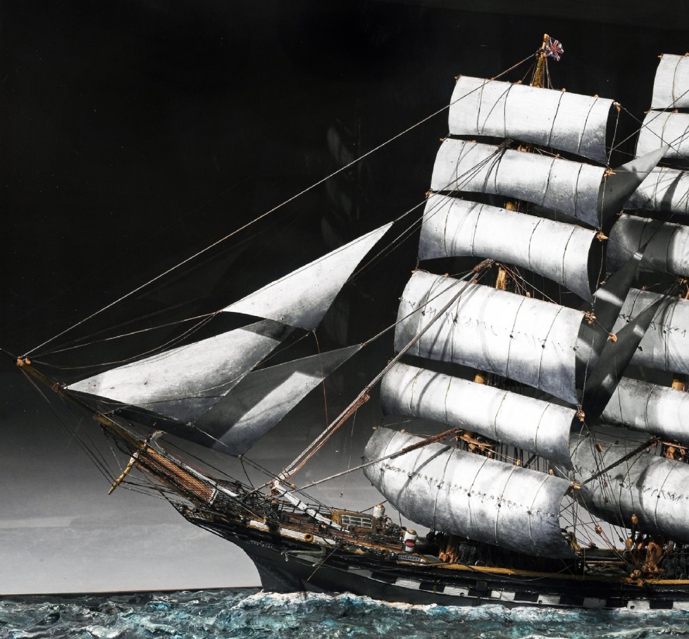 A detailed scale model of a three-masted tea clipper ship in full sail set within a seascaped display inside a glazed oak wood case with a brass plaque. View X