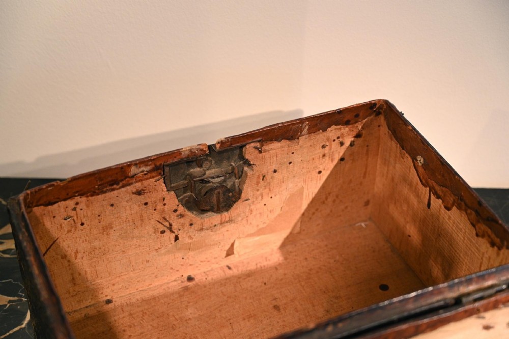 A 17th-century dark brown leather-bound travelling box with ornate pierced brass corner mounts, side latches, central lock plate, and a top carrying handle. View VI