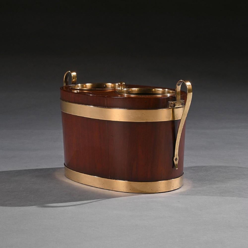 An oval George III mahogany table wine cooler featuring polished brass bands, two top bottle openings, a central finial, and elevated brass side handles. View II