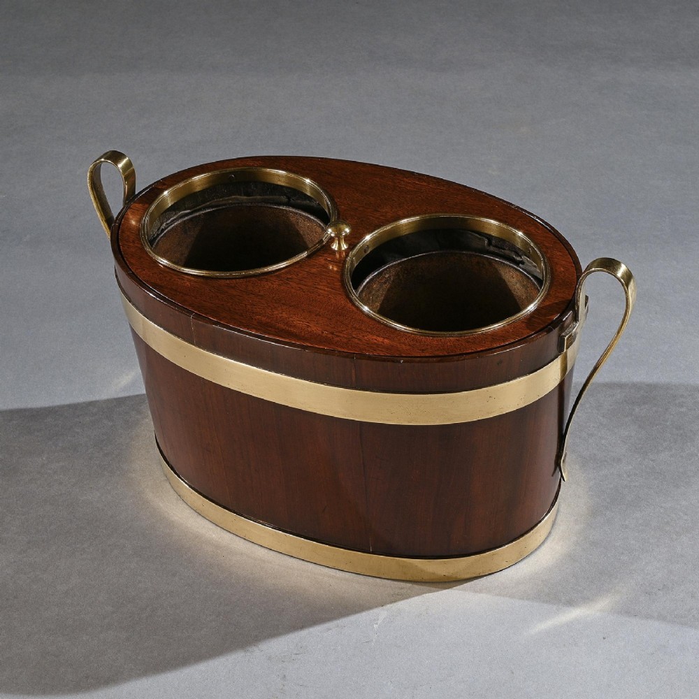 An oval George III mahogany table wine cooler featuring polished brass bands, two top bottle openings, a central finial, and elevated brass side handles. View V