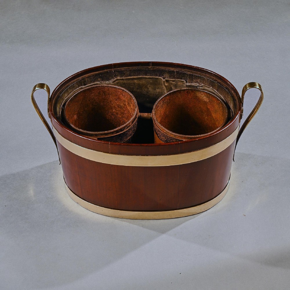 An oval George III mahogany table wine cooler featuring polished brass bands, two top bottle openings, a central finial, and elevated brass side handles. View VI
