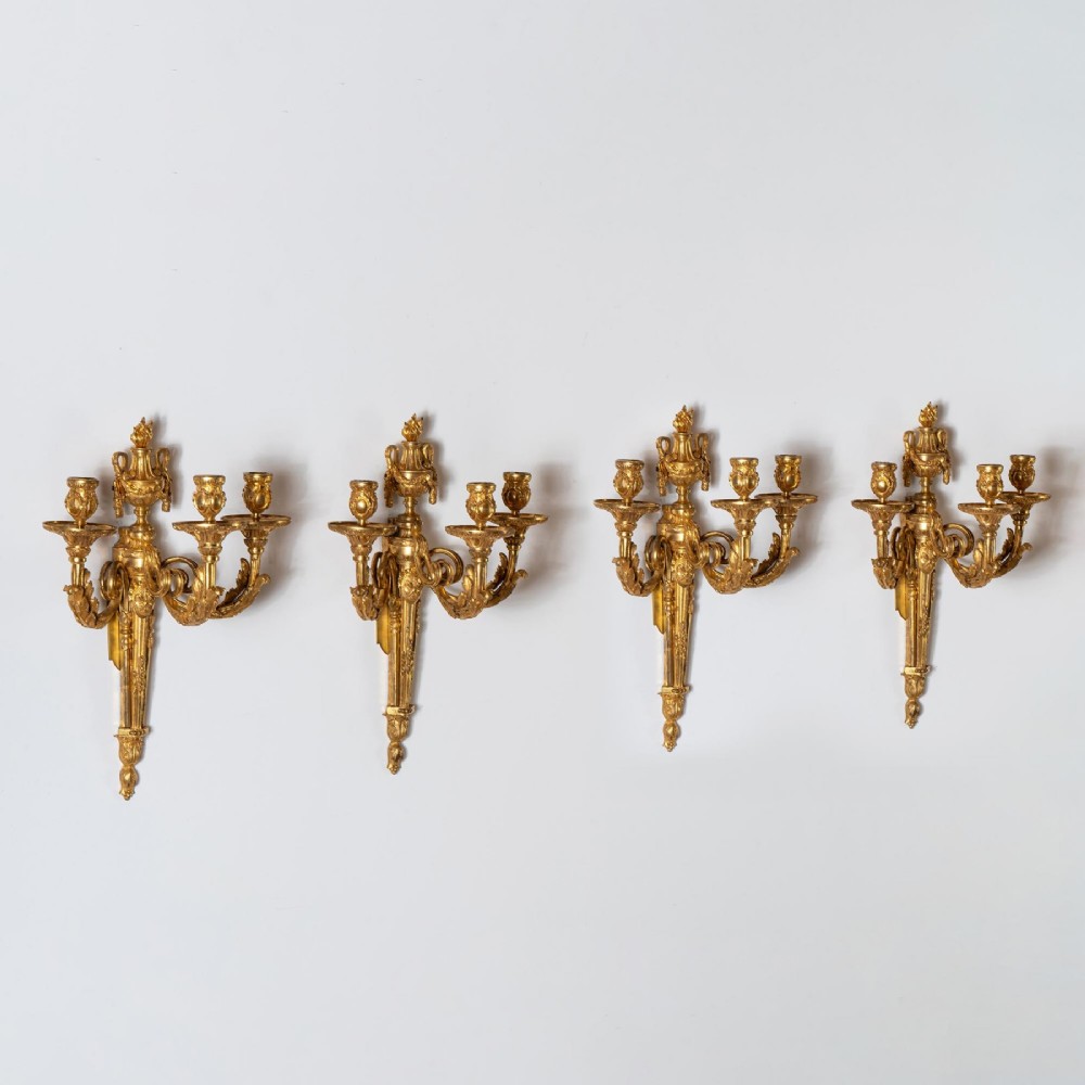 A set of four French Louis XV style gilded bronze wall lights, each featuring three scrolling foliate arms supporting candle sockets and an urn finial backplate. View II