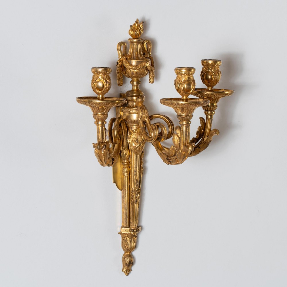A set of four French Louis XV style gilded bronze wall lights, each featuring three scrolling foliate arms supporting candle sockets and an urn finial backplate. View IX