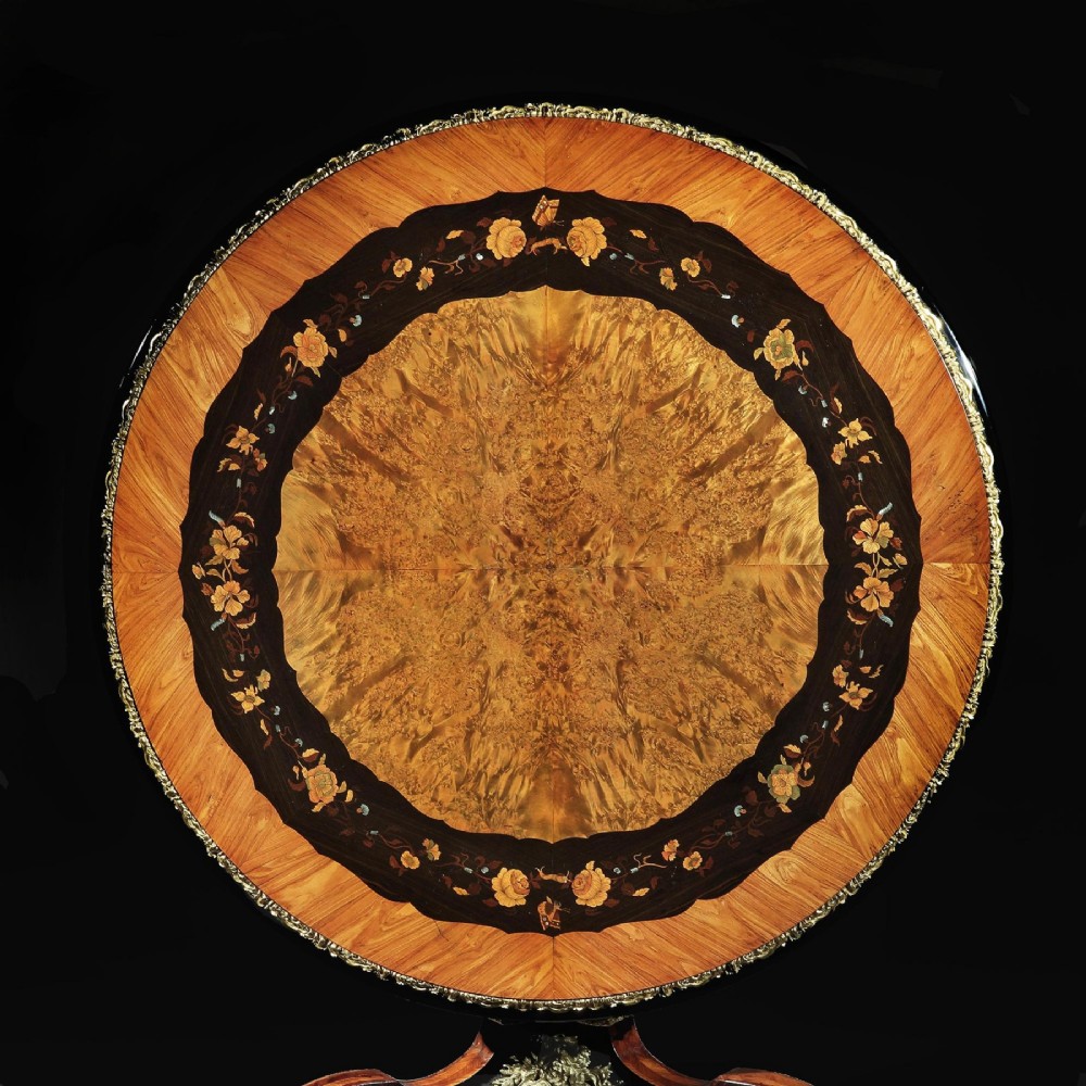 A 19th-century circular marquetry centre table featuring floral wood inlays, a tilt-top mechanism, and a dark carved tripod base with gilt-bronze mounts. View IV