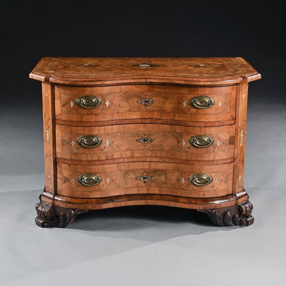 A mid-eighteenth-century German serpentine walnut commode featuring three drawers decorated with metal marquetry inlays, brass handles, and carved corner feet.