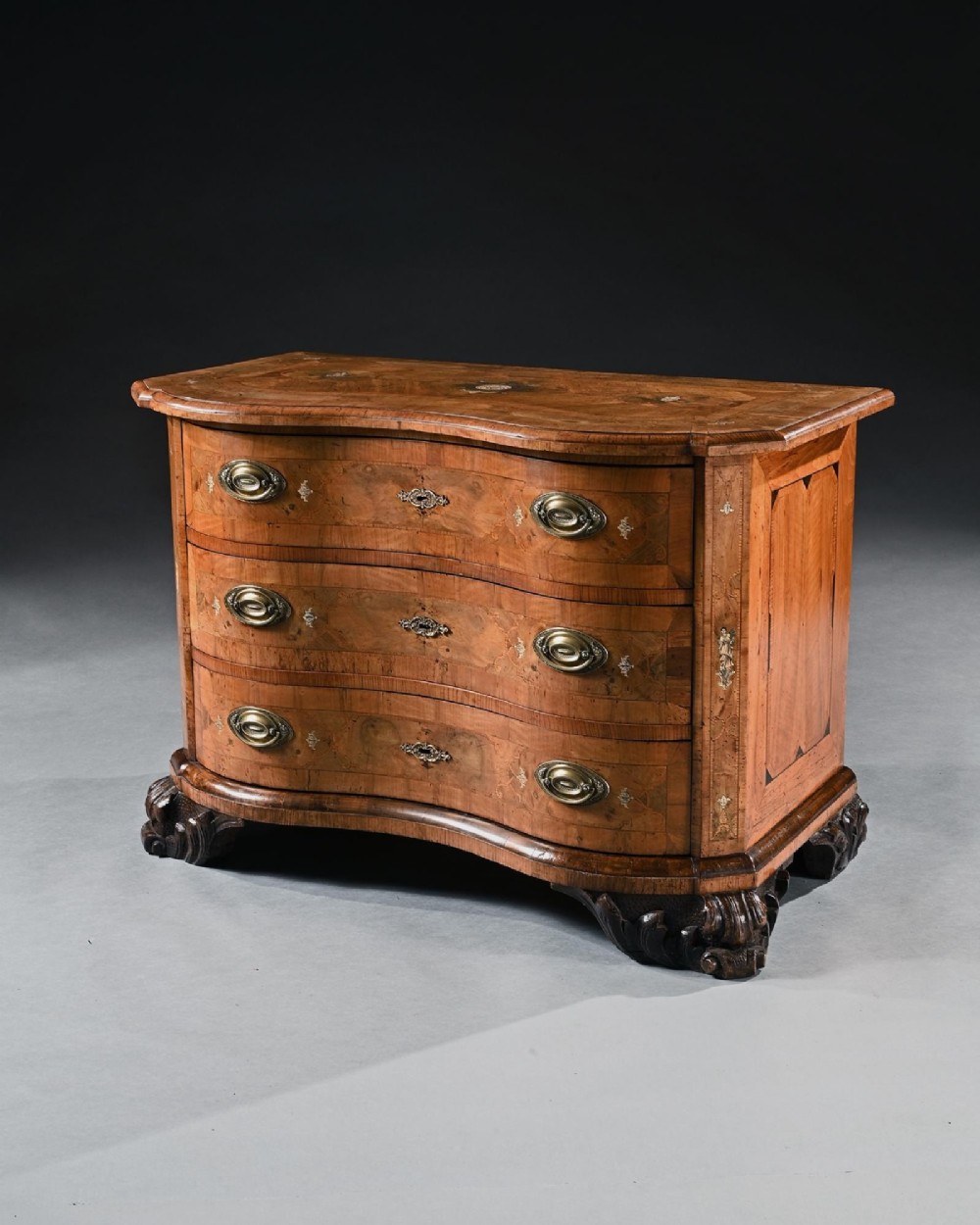A mid-eighteenth-century German serpentine walnut commode featuring three drawers decorated with metal marquetry inlays, brass handles, and carved corner feet. View IV