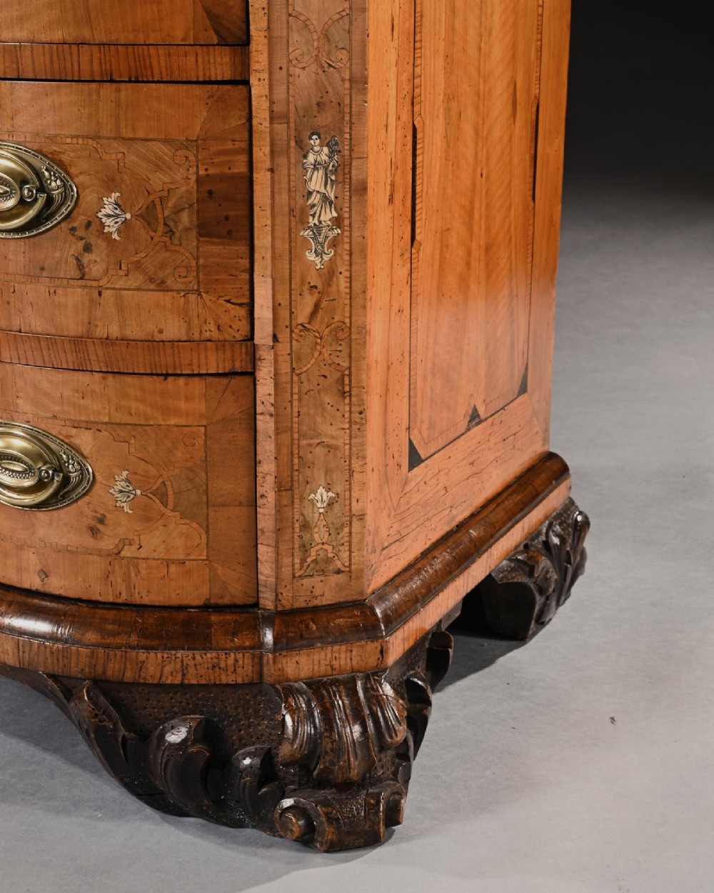 A mid-eighteenth-century German serpentine walnut commode featuring three drawers decorated with metal marquetry inlays, brass handles, and carved corner feet. View V
