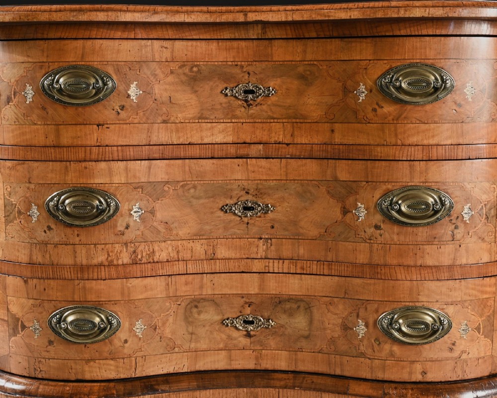 A mid-eighteenth-century German serpentine walnut commode featuring three drawers decorated with metal marquetry inlays, brass handles, and carved corner feet. View VI