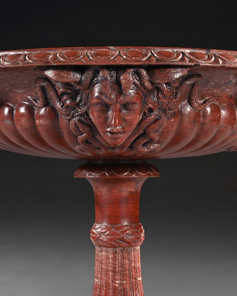 A carved deep red Rosso Antico marble tazza featuring serpent handles, a Medusa mask motif, and a fluted stem set upon a square pedestal base. View VI