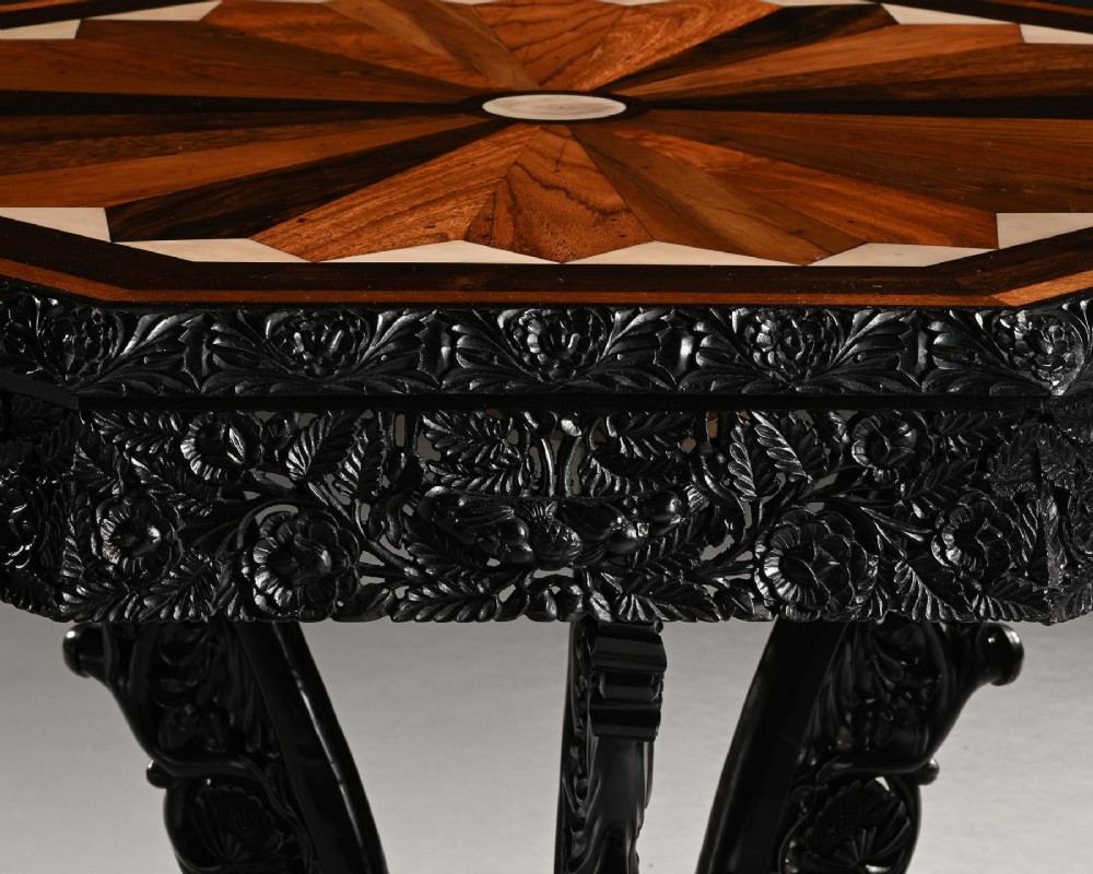 A 19th-century Ceylonese octagonal centre table crafted from dark ebony featuring a geometric starburst specimen wood inlaid top and deeply carved pedestal base. View VI
