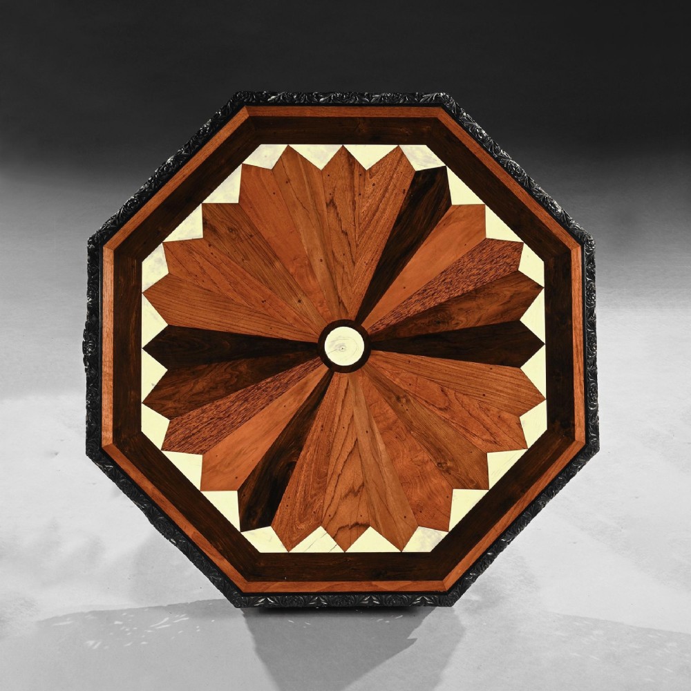 A 19th-century Ceylonese octagonal centre table crafted from dark ebony featuring a geometric starburst specimen wood inlaid top and deeply carved pedestal base. View VIII