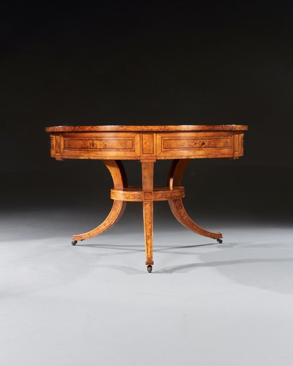 A circular early 19th-century Scandinavian drum table crafted from burr maple with frieze drawers, a lower shelf, and quadruped splayed legs with brass castors. View X