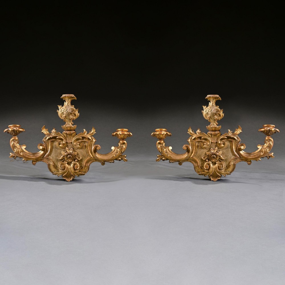 A pair of carved giltwood wall sconces featuring ornate rococo scrollwork, acanthus leaf motifs, and twin candle arms against a dark backdrop. View III