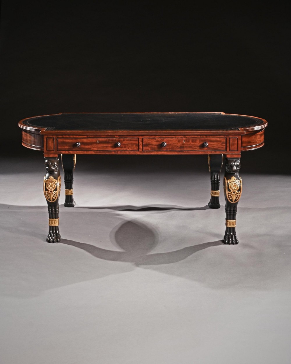 A Regency style mahogany writing table featuring a dark leather inset top, frieze drawers, and four ebonised monopodia legs ornamented with carved lion heads and gilt accents. View IV