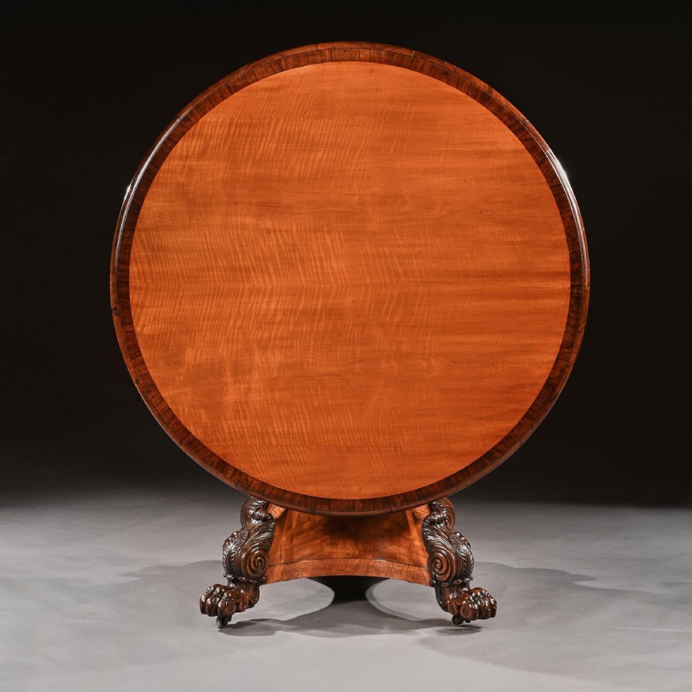 A circular 19th-century satinwood centre table with rosewood crossbanding, supported by a flared triangular pedestal base and carved scroll feet. View II