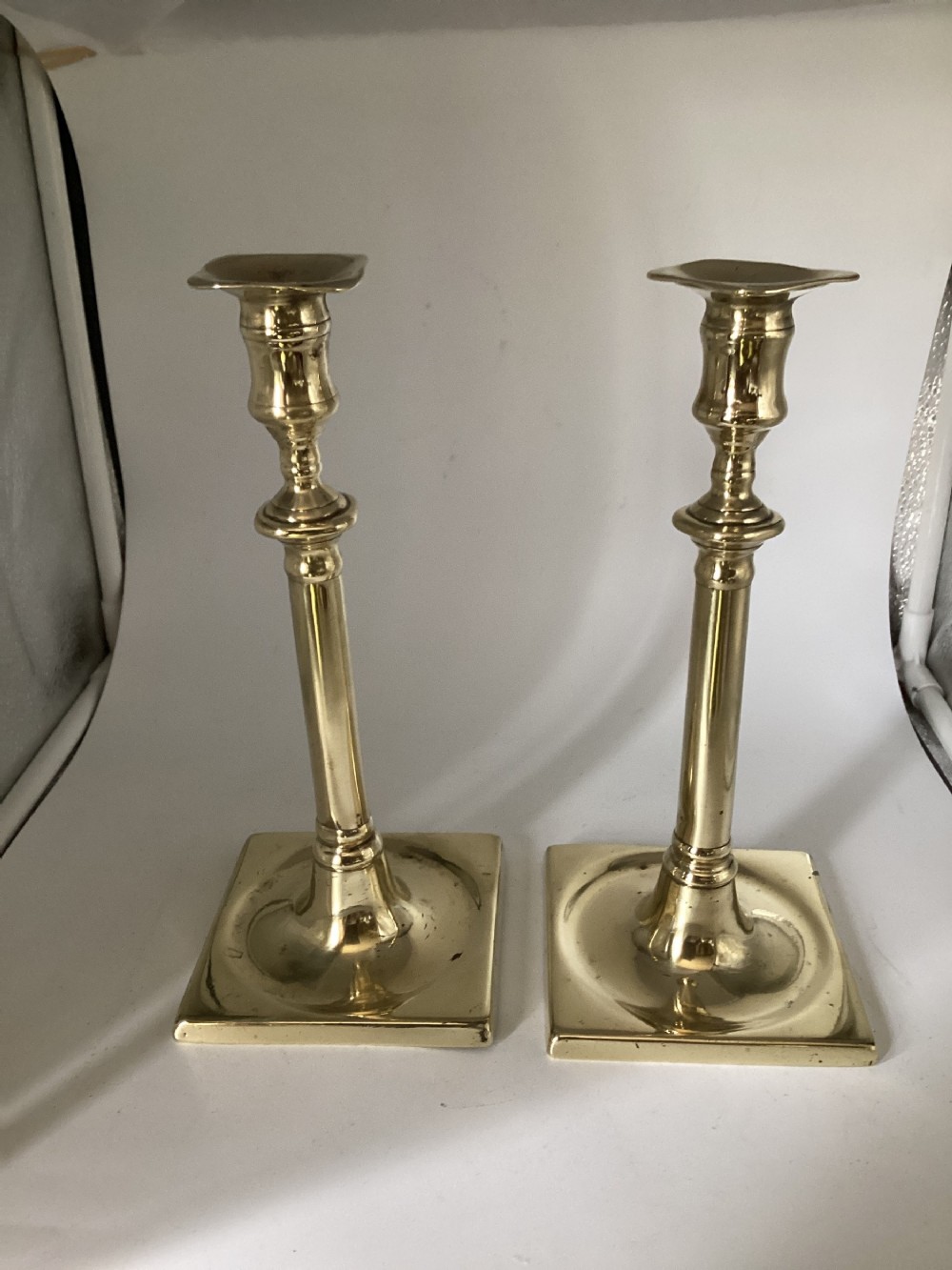 A pair of polished brass candlesticks featuring square recessed bases, turned stems with knopped details, and square top sconces.