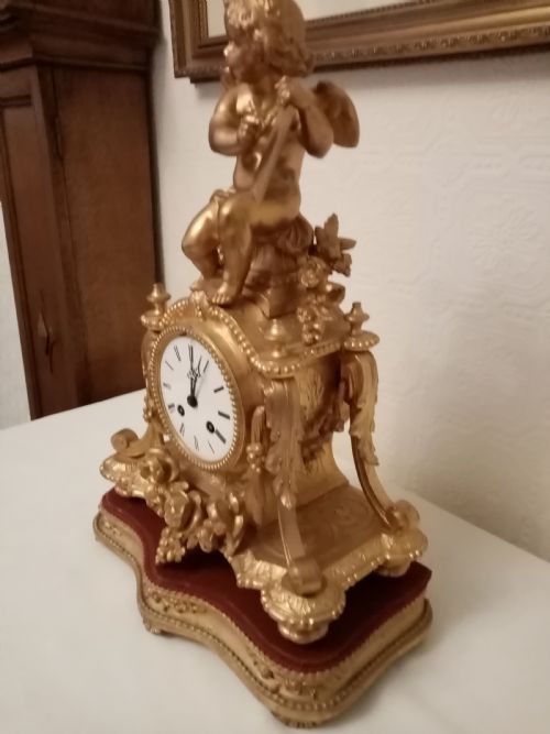 fig 1. An ornate gold gilt bronze mantel clock topped with a seated cherub figure writing on a scroll above an enamelled dial with Roman numerals.
