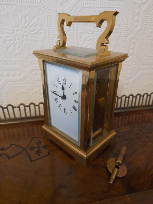fig 1. A rectangular brass carriage clock featuring clear glass side panels, a white enamel dial with black Roman numerals, and a shaped brass carrying handle.