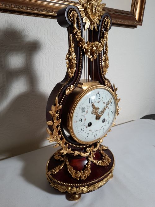 fig 1. A Louis XVI style lyre-shaped mantel clock crafted from dark mahogany with elaborate gilt bronze floral festoons, an Apollo sunburst finial, and a floral enamel dial.