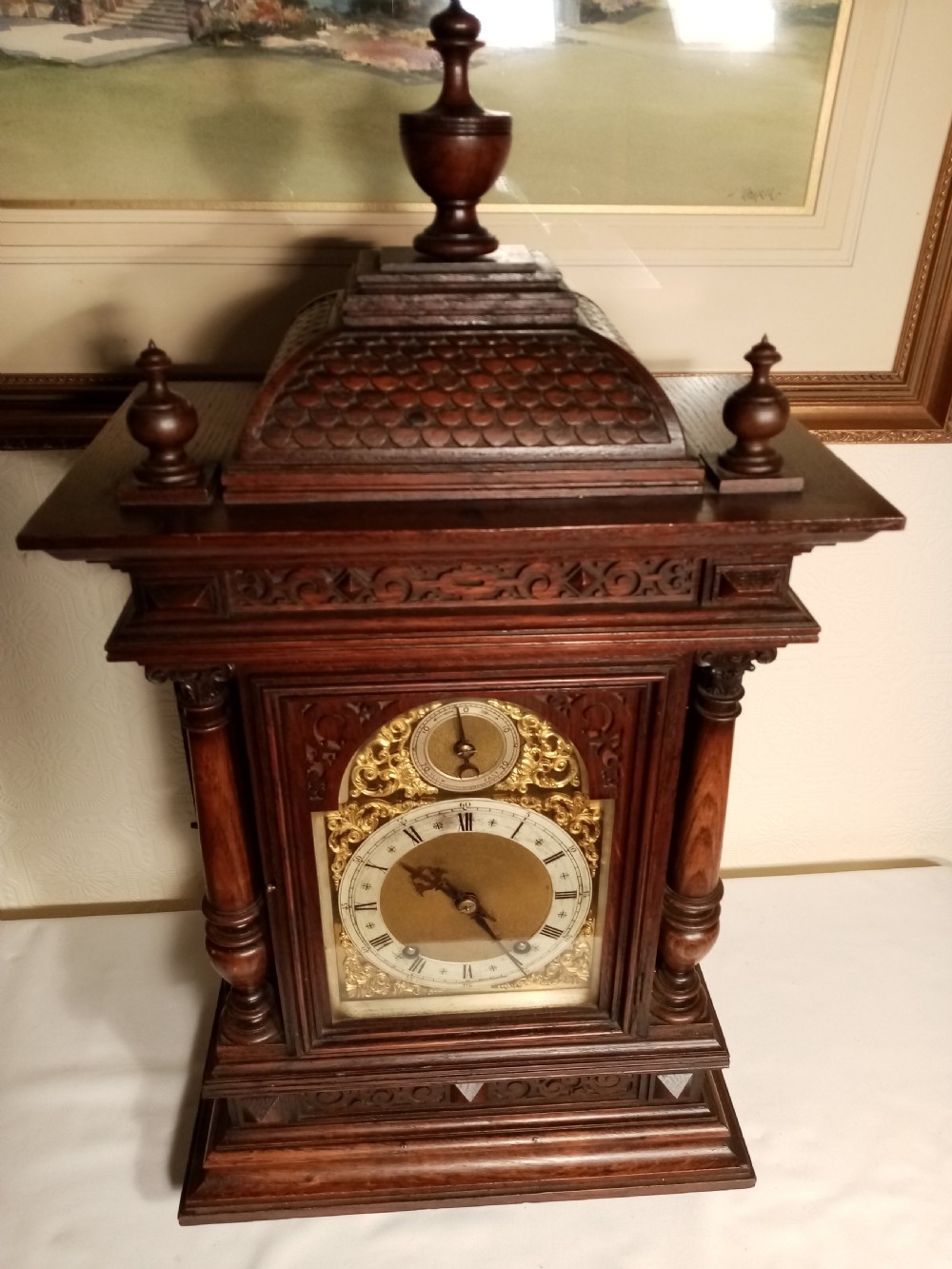 A large dark carved oak bracket clock featuring a decorative scale-patterned dome topped with a central finial, fluted side columns, and an ornate brass dial with a silvered chapter ring.