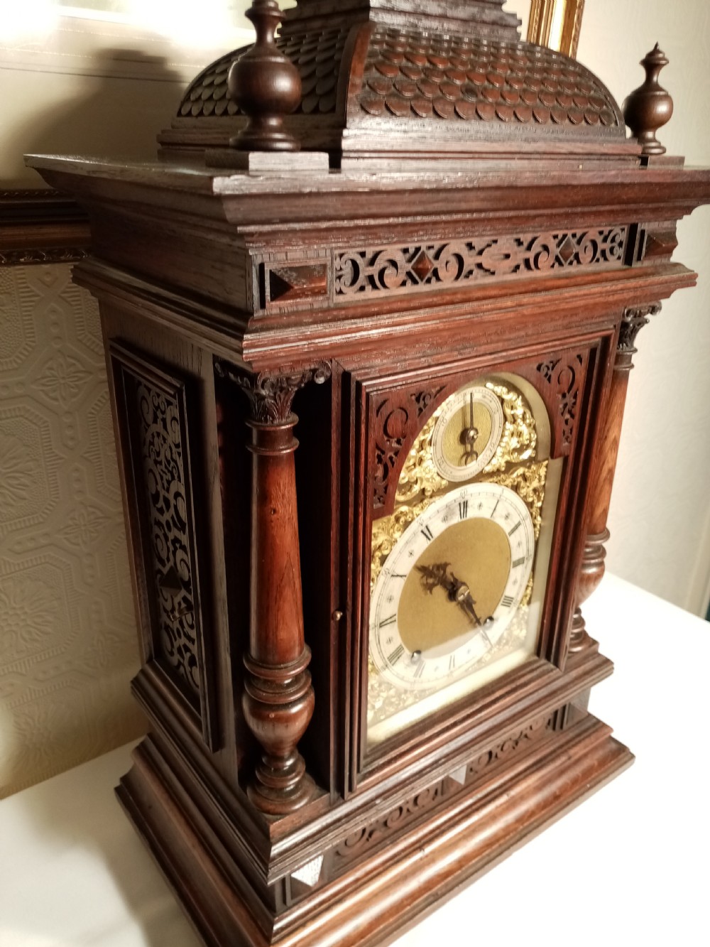 A large dark carved oak bracket clock featuring a decorative scale-patterned dome topped with a central finial, fluted side columns, and an ornate brass dial with a silvered chapter ring. View III