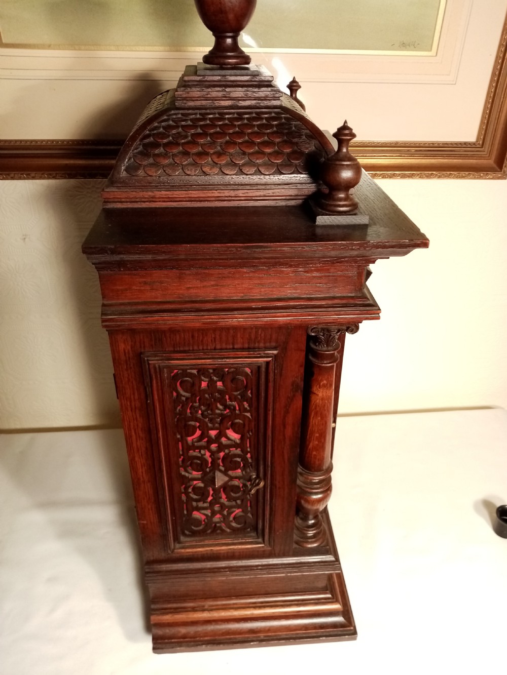 A large dark carved oak bracket clock featuring a decorative scale-patterned dome topped with a central finial, fluted side columns, and an ornate brass dial with a silvered chapter ring. View V
