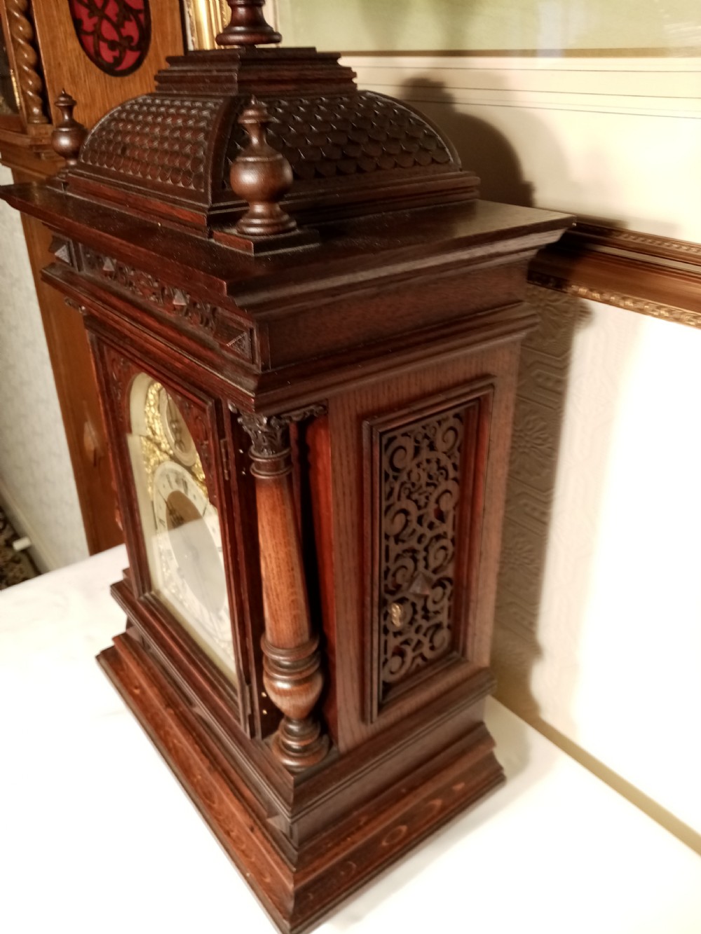 A large dark carved oak bracket clock featuring a decorative scale-patterned dome topped with a central finial, fluted side columns, and an ornate brass dial with a silvered chapter ring. View VI