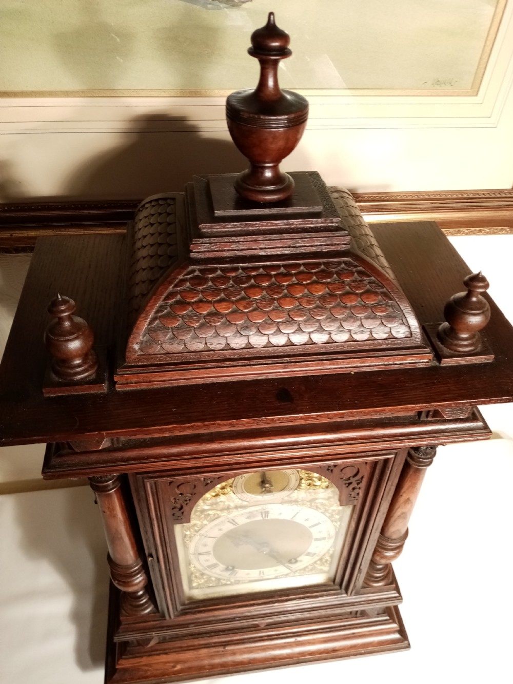 A large dark carved oak bracket clock featuring a decorative scale-patterned dome topped with a central finial, fluted side columns, and an ornate brass dial with a silvered chapter ring. View VII